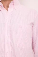 Camisa de corte clásico confeccionada en algodón, con diseño de rayas verticales finas en color rosa sobre fondo blanco. Presenta cuello abotonado, cierre frontal con botones, mangas largas con puños abotonados y un bolsillo en el pecho con el logo de la marca bordado.