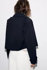 Trench corta de color negro con cuello de solapa, cierre frontal cruzado con botones y mangas largas con trabillas ajustables. Cuenta con bolsillos delanteros de vivo.