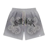 Bermuda de felpa color gris claro con efecto lavado (washed). Presenta un corte ancho con cintura elástica y cordón beige. El diseño frontal incluye un estampado serigrafiado en negro con texto y motivos florales/ornamentales en bordado blanco. Posee remaches metálicos cromados en los laterales y bolsillos.