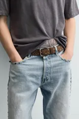 Cinturón de cuero gamuzado color marrón, con aplicación de tachas metálicas circulares y ojales decorativos. Cuenta con hebilla metálica de estilo envejecido y pasador a juego.