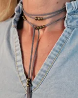 Collar estilo corbatín confeccionado en tiento de cuero gris, con diseño de doble vuelta y detalle de cuentas metálicas en color bronce.