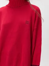 Pullover de punto color rojo, con cuello tortuga, corte holgado y mangas largas con puños acanalados en contraste con rayas blancas. Presenta un logo bordado en el pecho.