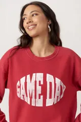 Sweater de tejido jaspeado color rojo, con cuello redondo y hombros caídos. Presenta un estampado frontal desgastado con la frase "GAME DAY" en tipografía deportiva.