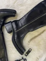 Bota alta tipo biker de cuero negro con cierre metálico lateral que recorre toda la caña. Presenta un diseño versátil con estribo y cadena metálica desmontables en la zona del tobillo, taco cuadrado y plataforma delantera.