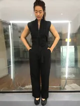 Mono largo de lino color beige, con detalle plisado en el frente, escote en V, sin mangas, lazo en la cintura, cierre invisible delantero y bolsillos laterales.