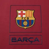 Camiseta deportiva color borgoña con cuello redondo y mangas cortas. Estampado en el pecho con el escudo del FC Barcelona y la palabra "Barça" en azul.