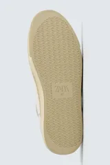 Zapatilla deportiva de estilo retro, color blanco con detalles en gris y negro. Presenta una suela de color beige en contraste y cordones blancos.