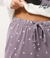 Pijama largo de dos piezas compuesto por una remera de manga larga color blanco con cuello redondo y estampa frontal de un gato con collar de perlas, y un pantalón color lila con estampado de lunares blancos, cintura elástica con cordón y puños ajustados en los tobillos.