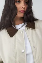 Campera acolchada corta color beige, con cuello de pana en contraste color marrón. Cuenta con cierre frontal de cremallera y botones a presión, bolsillos laterales, y puños y bajo elásticos.