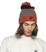 Gorro de lana tipo beanie con pompón superior, diseño de franjas horizontales en colores terracota, gris y negro. Incluye un parche circular con logo en la parte frontal.
