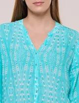 Blusa turquesa de silueta suelta confeccionada en tejido liviano, con escote en V, pequeña cartera frontal con botones decorativos y mangas 3/4 con presilla para arremangar. Presenta patrón estampado geométrico tipo tie-dye y ruedo curvo.