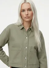 Chaqueta corta de algodón color verde oliva, con cuello camisero, cierre frontal de botones metálicos y dos bolsillos de parche en el frente. Presenta un corte relajado.