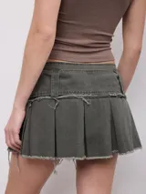 Minifalda plisada color tostado con diseño de talle bajo y bordes deshilachados. Incluye un cinturón ancho incorporado con hebilla metálica rectangular y detalles de tachas.