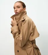 Trench coat largo de Marfinno, confeccionado en gabardina de poliéster. Presenta cuello con solapas, aletas en los hombros, cierre frontal con botones y cinta para lazo en la cintura.