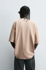 Camiseta de algodón color beige, corte relaxed fit, con cuello redondo y manga corta. Presenta un estampado gráfico grande en negro con motivos variados como una estación de servicio antigua, un caballo alado, una polilla, un pájaro y el texto "RUSTED ROSE".