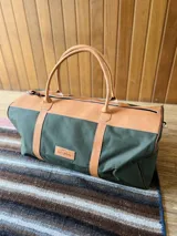 Bolso de viaje tipo duffel de lona negra con detalles en cuero tostado, asas de mano y correa ajustable para el hombro.