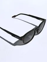 Lentes de sol con armazón de pasta color negro mate y diseño de marco redondeado. Presentan detalles en color rojo en las patillas y el frente, con el logo de la marca grabado en el lateral.