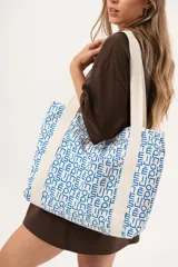 Bolso tipo tote de gran capacidad, confeccionado en tela con estampado geométrico de letras y formas en azul sobre fondo blanco. Cuenta con correas anchas de lona en color crudo, cierre principal y bolsillo delantero.