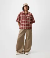 Camisa de franela de algodón con diseño a cuadros en tonos rojo y beige, de corte holgado relaxed, mangas cortas, cuello tradicional y cierre frontal con cremallera. Cuenta con un bolsillo aplicado en el pecho con detalle de botón metálico.