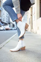 Bota tipo texana de caña baja en color blanco, con taco cuadrado de madera y punta fina. Presenta costuras decorativas en el lateral y un diseño clásico de estilo western.