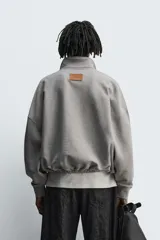 Sudadera de corte boxy fit en color gris, confeccionada en tejido de algodón con interior afelpado. Presenta cuello solapa, cierre frontal con botones a presión, manga larga con puños acanalados y bolsillo frontal tipo canguro. El bajo cuenta con un acabado elástico.