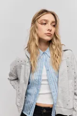 Campera gris melange de calce relajado, con capucha, cierre frontal y bolsillos laterales. Presenta puños y cintura de rib, y detalles de tachas metálicas en las mangas y la parte frontal.