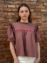Blusa color chocolate con mangas cortas abullonadas, cuello redondo con volados y detalles de costura en zigzag de color rojo en el pecho y volados oscuros en los hombros.