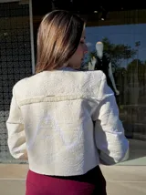 Chaqueta corta de estilo estructurado en color beige, con diseño de brocado floral en relieve y terminaciones desflecadas en el frente y cuello. Presenta mangas largas y cierre frontal oculto.