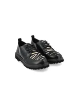 Zapato acordonado de cuero negro con detalle de estampado animal print tipo cebra en la lengüeta y empeine. Presenta suela track dentada con costuras amarillas visibles.