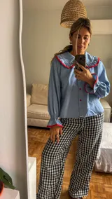 Camisa de color celeste con cuello bobo grande y puños con volados, ambos con ribetes en color rojo. Presenta cierre frontal con botones rojos.