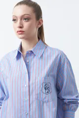 Camisa a rayas verticales blancas y marrones, de manga larga con puños abotonados, cuello camisero y botones frontales. Incluye un bolsillo en el pecho con bordado sutil.