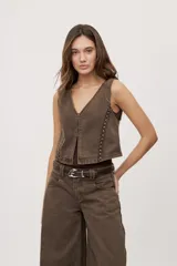 Chaleco corto de denim marrón con escote en V y diseño sin mangas. Presenta terminación lavada y detalles laterales con entramado y tachas metálicas.