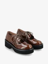 Zapato tipo guillermina con cordones, confeccionado en cuero negro con detalles de perforaciones estilo brogue. Presenta una suela de plataforma gruesa y tacón cuadrado.