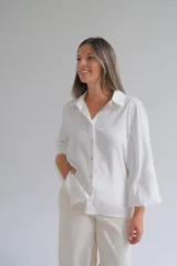 Camisa blanca de corte holgado con cuello clásico, cierre frontal de botones y mangas tres cuartos con sutil detalle bordado en los puños.