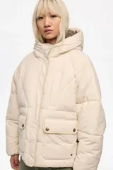 Chaqueta acolchada tipo puffer de corte globo en color beige, con cuello subido, capucha, cierre frontal con cremallera y botones de presión, y bolsillos delanteros con tapeta.