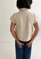 Top de algodón color crema, de manga corta y cuello alto estructurado. Cuenta con cierre frontal cruzado con botones metálicos a presión y costuras decorativas.
