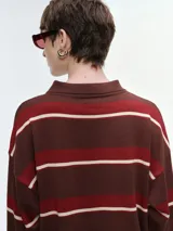 Buzo tejido de punto a rayas horizontales en tonos marrón, rojo y crema, con cuello polo y cierre frontal de botones. Presenta mangas largas con puños elastizados y un corte holgado.