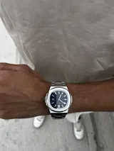 Reloj de pulsera con diseño inspirado en el modelo Nautilus, con caja y malla de acero inoxidable. Presenta una esfera azul con textura estriada horizontal, índices aplicados luminiscentes y ventana de fecha a las tres. Incluye cierre desplegable tipo mariposa.