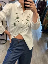 Camisa de algodón color marrón con cuello clásico y cierre frontal de botones. Presenta mangas largas con puños elásticos y detalle de bordados florales en la parte frontal.