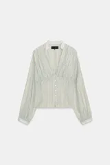 Camisa de manga larga con cuello redondo y escote en pico. Presenta un diseño a rayas verticales en tonos marrones y celestes, con detalle de pliegues en el delantero y los hombros. Cierre frontal con botones y puños ajustados.