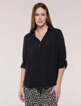Blusa holgada de color negro con cuello camisero, escote en V y cierre parcial de tres botones metálicos. Tiene mangas largas convertibles a tres cuartos con presilla y acabado en crepé.