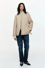Campera acolchada de nylon beige con diseño guateado, cuello redondo y cierre frontal. Cuenta con hombros caídos, mangas largas, bolsillos laterales y cordón de ajuste en la cintura con tiras colgantes.