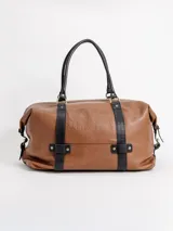 Bolso de viaje grande de cuero marrón con detalles en cuero negro, doble asa de mano y tiras decorativas verticales.