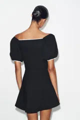 Vestido corto de tejido bouclé en color negro, con escote recto y mangas cortas abullonadas. Presenta detalles de vivos en color blanco a contraste en el escote, el centro del delantero y en los falsos bolsillos frontales.