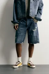 Bermuda baggy fit confeccionada en denim de algodón con lavado azul y efecto rotos en toda la prenda. Presenta diseño de cinco bolsillos, detalles de costuras en las perneras, bajo acabado irregular y cierre frontal con cremallera y botón.