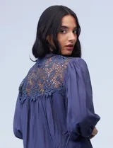 Blusa color beige de gasa con cuello a la base, escote en V y abertura central con botones. Presenta detalles de crochet en hombros y espalda, mangas 3/4 amplias con puño y botón. El largo de la espalda es mayor al delantero.