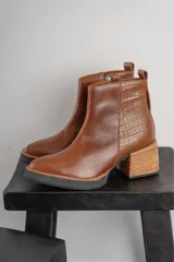 Bota corta de cuero negro con acabado combinado mate y charol. Presenta una tira decorativa con tachas metálicas y hebilla en el tobillo, tacón cuadrado robusto y suela con plataforma.