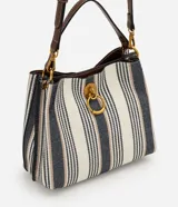 Cartera bucket pequeña de Satinato con diseño de rayas verticales en tonos blanco, azul marino y beige, confeccionada en tejido estructurado. Cuenta con asa de mano fija, asa larga desmontable de cuero sintético marrón, detalles de herrajes metálicos dorados, argolla metálica colgante en el frente y cierre superior.