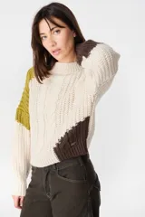 Sweater de punto con diseño de bloques de color en tonos crema y marrón. Presenta cuello redondo, mangas largas y una silueta holgada con detalle de placa metálica decorativa en el lateral.