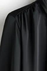 Blusa de gasa negra con cuello alto y lazada frontal, mangas largas abullonadas con puños abotonados y detalle de frunces en los hombros.